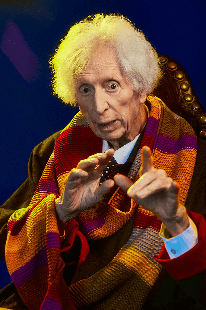 Tom Baker sentado en una silla con una réplica de su bufanda del Cuarto Doctor con las manos extendidas