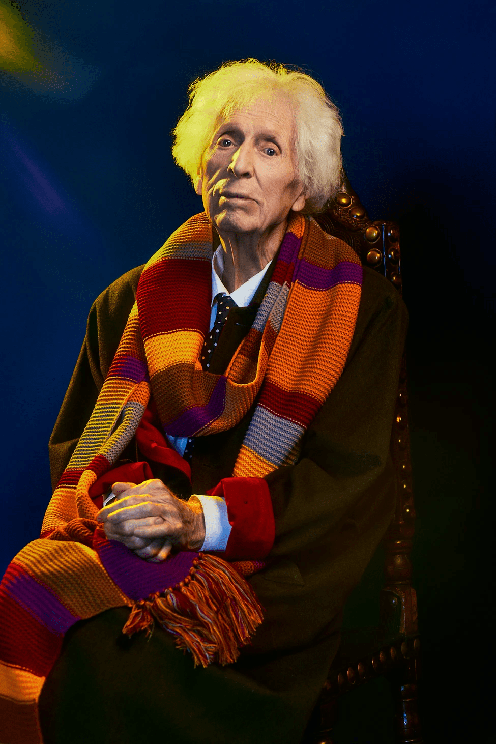 Tom Baker sentado en una silla con una réplica de su bufanda del Cuarto Doctor